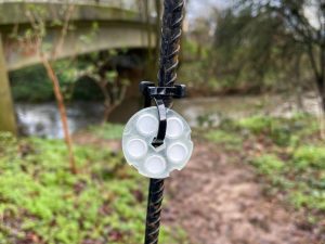 Close-up of a plastic cap secured to a thin pole with a cable tie in front of a river with a bridge and greenery in the background.